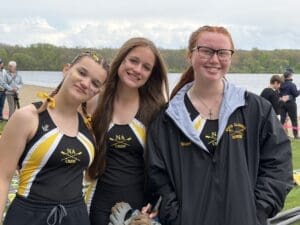 Three young female rowers pose for a photo by a lake, wearing matching NA Crew uniforms, smiling and cheerfully decorated.