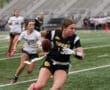 Female flag football player in black jersey runs with the ball while another player chases her on the field.
