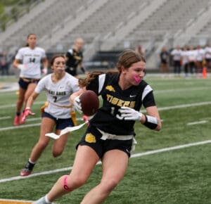 Female flag football player in black jersey runs with the ball while another player chases her on the field.