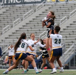 Girl catching a football during a competitive flag football game, with teammates in action on a field.