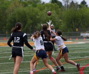 Girls competing in flag football, with players reaching for a pass while a referee oversees the game.