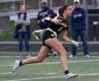 Female athlete in action, catching a football during a game on a field with spectators in the background.