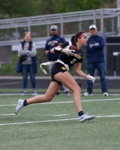 Female athlete in action, catching a football during a game on a field with spectators in the background.