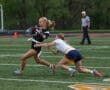 Female athletes competing in flag football; one player evades a defender while carrying the ball on the field.