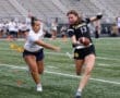 Two athletes compete in women's flag football on a grassy field, one dodging a tag while holding a football.