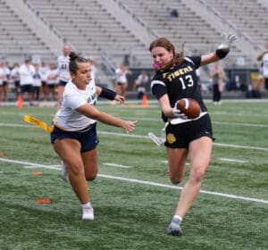 Two athletes compete in women's flag football on a grassy field, one dodging a tag while holding a football.