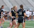 Cheerful girls in black jerseys celebrate during a flag football game on a sports field with bleachers in the background.