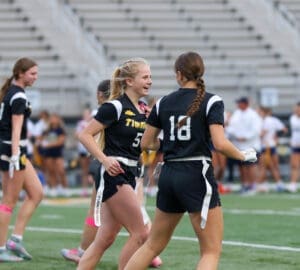Cheerful girls in black jerseys celebrate during a flag football game on a sports field with bleachers in the background.