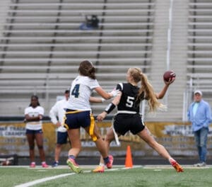 Girl in black sports uniform throws football while being defended by an opponent in a women's flag football game.