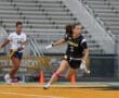 Girl in black Tigers jersey sprinting on a field, playing lacrosse, with a player in white behind her.