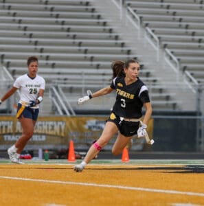 Girl in black Tigers jersey sprinting on a field, playing lacrosse, with a player in white behind her.