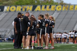 Girls' flag football team in black uniforms huddles with a coach on the field at North Allegheny High School.