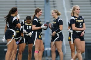Girl's flag football team in black jerseys smiles and prepares for a game, showcasing teamwork and athletic spirit.