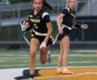 Two female athletes in black and yellow football jerseys play on an outdoor field, one running with a football.