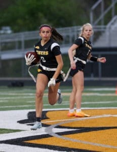 Two female athletes in black and yellow football jerseys play on an outdoor field, one running with a football.