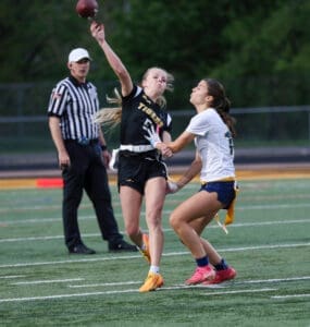 Female flag football player throws a pass while being defended by an opponent during a game, with an official observing.