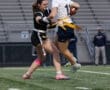 Two female athletes compete in a flag football game, showcasing action on a field with spectators in the background.