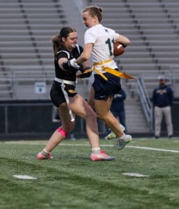 Two female athletes compete in a flag football game, showcasing action on a field with spectators in the background.