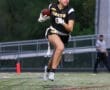 Female football player in a black jersey running with a football on a field during an evening game.