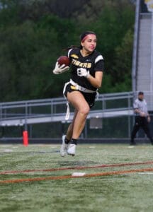 Female football player in a black jersey running with a football on a field during an evening game.