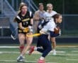 Two female athletes compete in a flag football game, one evading the other on a grassy field. Team colors are visible.