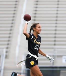 Girl athlete in a black football uniform, smiling and preparing to throw a football on a field with bleachers in the background.