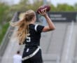 Girl in a black jersey throws a football, showcasing athleticism on a sports field with stadium seating in the background.