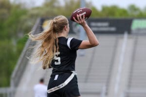 Girl in a black jersey throws a football, showcasing athleticism on a sports field with stadium seating in the background.