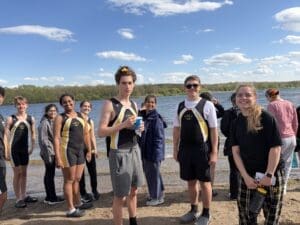 A group of young rowers in team uniforms enjoy a sunny day by the river during training or a competition.
