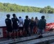 Group of young people standing by a lake, enjoying nature on a sunny day. Wooden benches in the foreground.