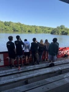 Group of young people standing by a lake, enjoying nature on a sunny day. Wooden benches in the foreground.