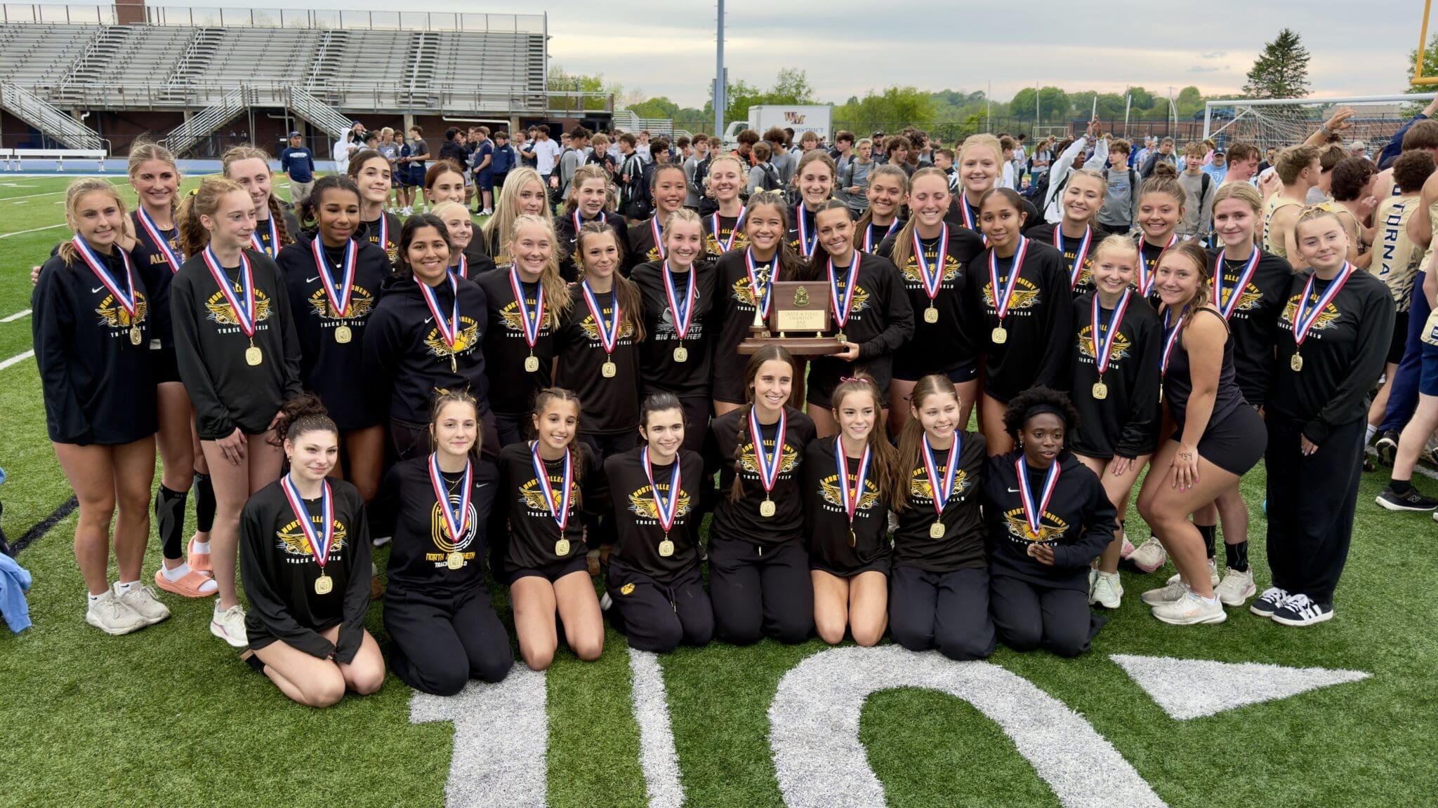 High school track team celebrates victory with medals and a trophy on the field, showcasing teamwork and achievement.
