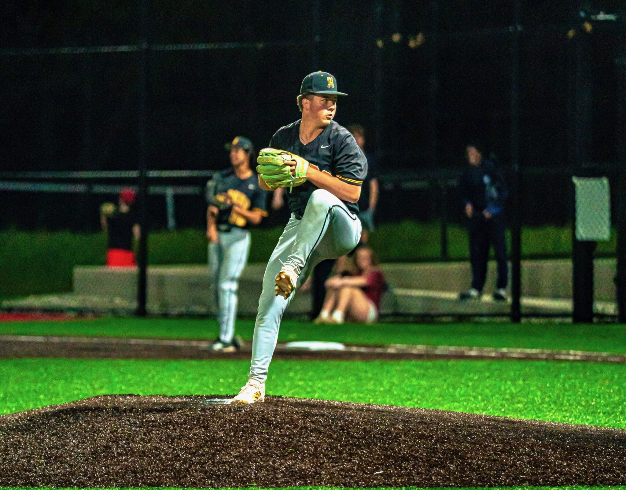 Baseball pitcher winding up on the mound under stadium lights, with teammates and spectators in the background.