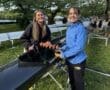 Two young women smiling while preparing a rowing boat at a scenic outdoor location, surrounded by greenery.