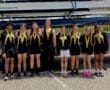 Group of eight young rowers celebrating with medals, standing in front of their boat trailer on a sunny day.