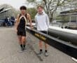 Two young rowers standing beside a boat labeled Landau Cruiser at a rowing event, showcasing teamwork and athleticism.