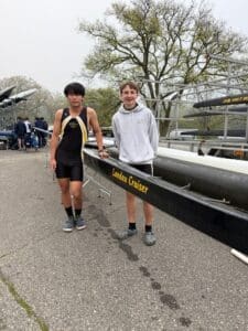 Two young rowers standing beside a boat labeled Landau Cruiser at a rowing event, showcasing teamwork and athleticism.