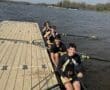 Rowing team members smiling on their boat, ready for practice on a sunny day by the water.