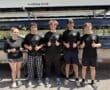 Five young rowers in matching black shirts giving thumbs up, standing by their rowing boat and smiling outdoors.