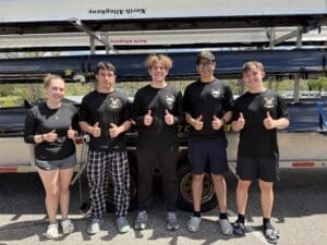 Five young rowers in matching black shirts giving thumbs up, standing by their rowing boat and smiling outdoors.
