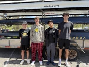 Four young rowers pose together by their boat trailer, showcasing their team spirit on a sunny day.