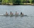 Four rowers in a boat compete on a serene river, showcasing teamwork and athleticism on a sunny day.