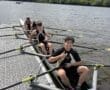 Teenage rowers preparing for practice on a river, with a city skyline in the background and cloudy skies above.