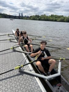 Teenage rowers preparing for practice on a river, with a city skyline in the background and cloudy skies above.