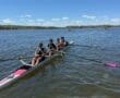 Four young rowers in a boat rowing on a calm lake under a clear blue sky, with clouds in the background.