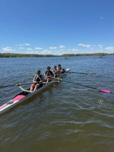 Four young rowers in a boat rowing on a calm lake under a clear blue sky, with clouds in the background.