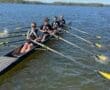 A rowing team of five athletes in a shell on a calm lake under a clear blue sky, showcasing teamwork and sport.