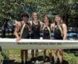 Four rowers in black and yellow gear pose together by their boat, celebrating camaraderie and teamwork at a sunny event.