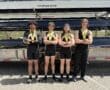 Four rowers proudly display medals, posing confidently in athletic gear by their boat trailer on a sunny day.