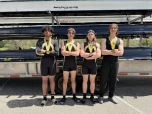 Four rowers proudly display medals, posing confidently in athletic gear by their boat trailer on a sunny day.